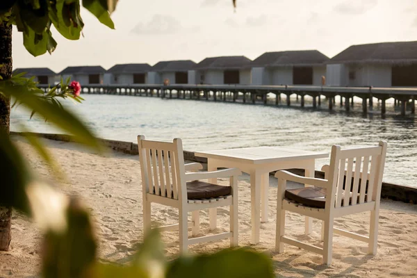 Mesa y sillas vacías blancas en la playa tropical resrraunt en maldivas. Laguna azul del océano y bungalows de agua en el fondo. No hay gente . —  Fotos de Stock