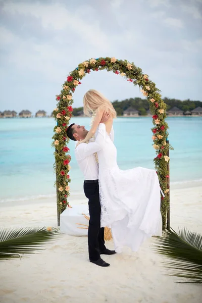 Happy groom holds bride on his hands under wedding ceremony arch on tropical island beach on Maldives. Turquoise ocean lagoon, white sand and bungalows of luxury spa resort on background. Honeymoon.