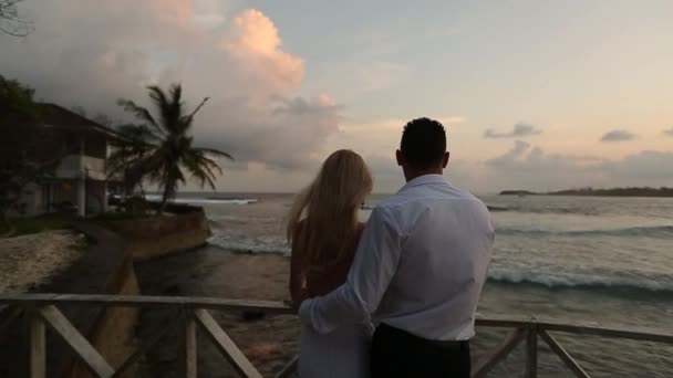 Vista trasera de la novia y el novio disfrutando de la puesta de sol en la playa tropical cerca de la balaustrada de vacaciones. Recién casados abrazando y viendo el océano después de la ceremonia en la luna de miel. Boda tropical en Maldivas spa de lujo — Vídeos de Stock