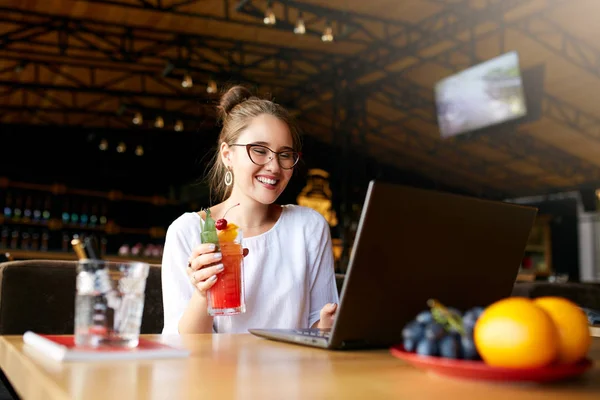 Smiling mixed race woman with cocktail in hand works with laptop. Businesswoman in glasses drinks juice for body hydration while working. Attractive designer quench thirst. Healthy lifestyle theme. — Stock Photo, Image