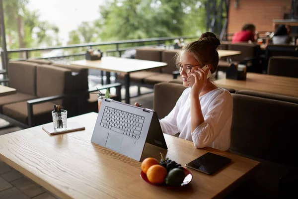 Mixed race freelancer working with convertible laptop and talking on cellphone with client in cafe. Asian caucasian businesswoman conducts negotiations via phone call. Multitasking business concept.