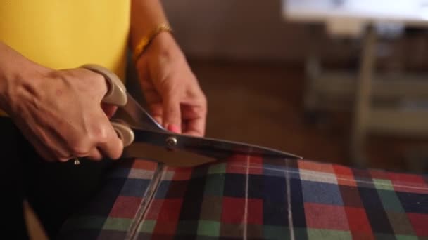 Vista cercana de las manos de mujer caucásica corte de tela con tijeras en el estudio de diseño. Tijeras de costurera tela a cuadros. La mujer crea ropa de diseño. Máquinas de coser en segundo plano. Mesa de corte . — Vídeos de Stock