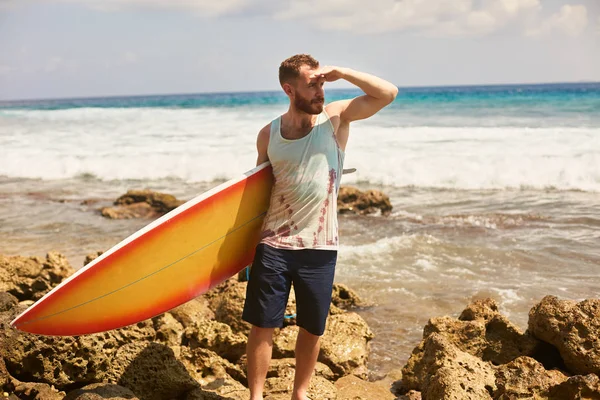 Surfista barbuto con una tavola da surf sta camminando sulla spiaggia e guardando le onde sul posto — Foto Stock