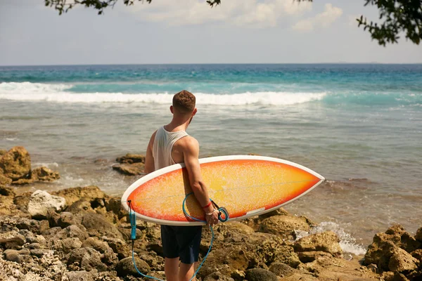 Surfista barbuto con una tavola da surf sta camminando sulla spiaggia e guardando le onde sul posto — Foto Stock