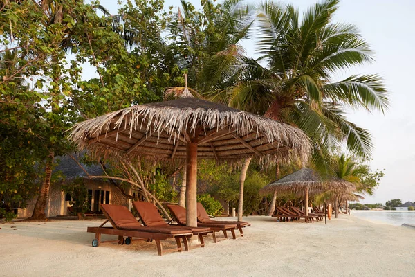 Beach deck chairs under straw umbrellas. Indian ocean coastline on Maldives island. White sandy beach and calm sea. Travel and vacation concept. — Stock Photo, Image