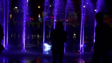 Geceleri renkli dans eden çeşme. Su jetleri siyah koyu arka plan üzerine ağır çekim portre görüntüleri. Tanınmayan insanlar siluetleri.