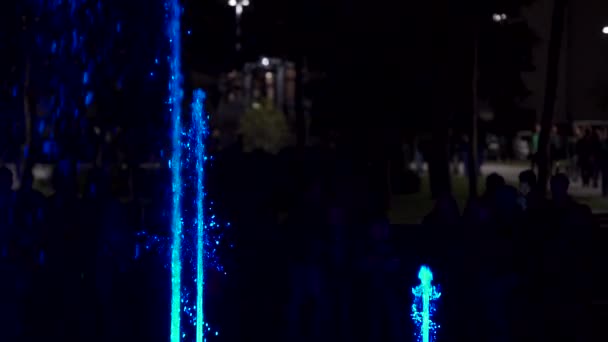 Kleurrijke dansende fontein in de nacht. Slow-motion close-up beelden van stralen water op een donkere achtergrond. Onherkenbaar mensen silhouetten. — Stockvideo