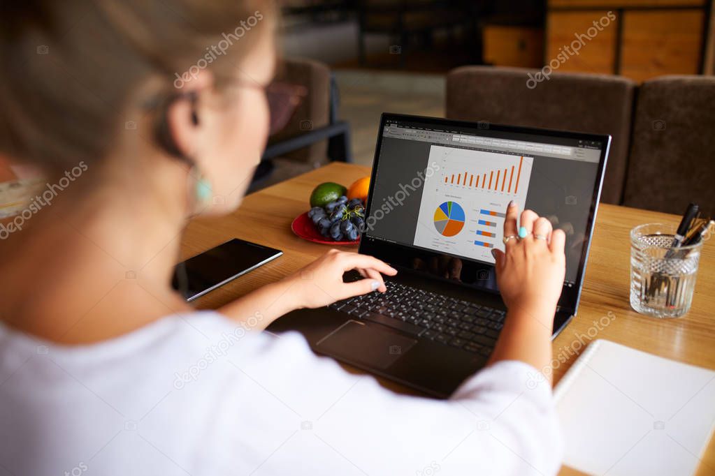 Back view of young business or student woman working at cafe with laptop computer, using touchscreen with finger and editing presentation document with charts and diagrams. Telecommuting concept.