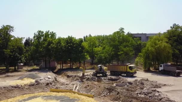 Yellow excavator digs ground of foundation pit and handling cargo with a bucket to the truck. Groundwork preparation at fountain construction site. 4k aerial footage of park reconstruction. — Stock Video