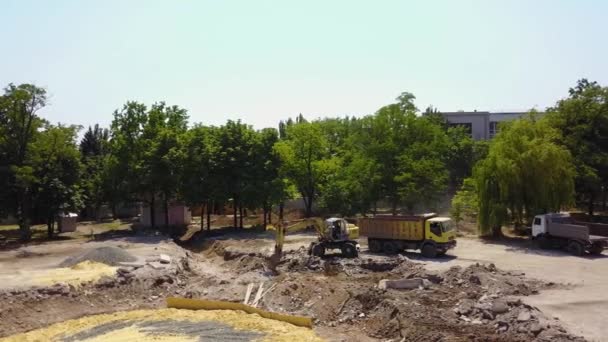 A escavadora amarela cava o solo do poço de fundação e manuseia a carga com um balde para o caminhão. Preparação de terraplenagem no canteiro de obras da fonte. 4k imagens aéreas de reconstrução do parque . — Vídeo de Stock