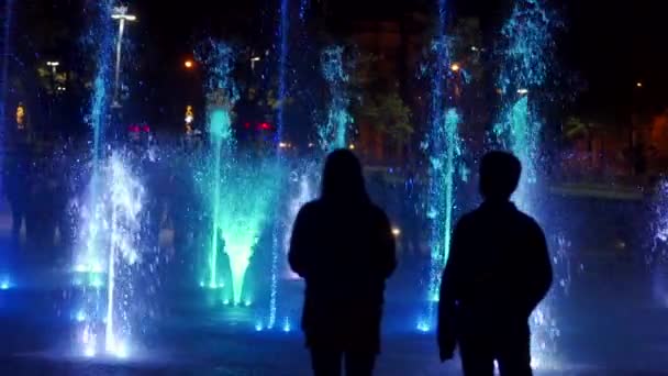 Colorida fuente de baile por la noche. Imágenes de primer plano en cámara lenta de chorros de agua sobre un fondo oscuro. Siluetas de personas irreconocibles . — Vídeo de stock