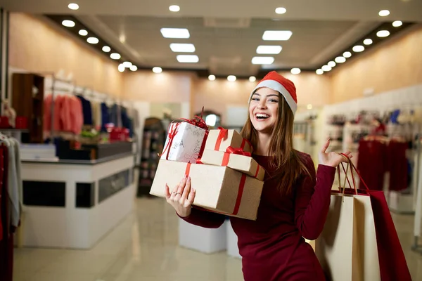 Happy girl shopping gifts in mall on christmas sale. New year holidays shopping idea concept. Smiling woman with colorful paper presents bags and gift boxes wearing christmas hat in store or shop. — Stock Photo, Image