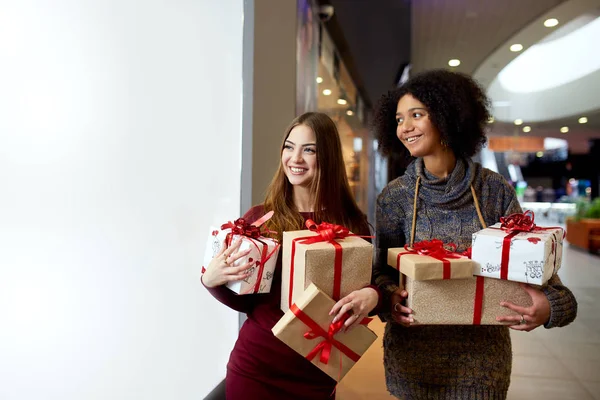 Two mixed race women with gift boxes in hands near storewindow. Multi ethnic girls smiling with presents on christmas new year sale. Caucasian and african american females buy presents for holidays. — Stock Photo, Image