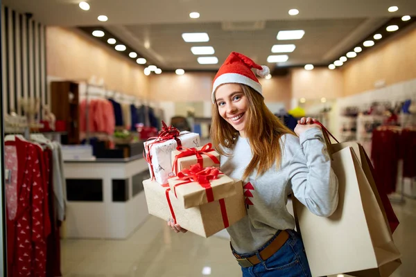 Joyeux cadeaux shopping fille dans le centre commercial sur la vente de Noël. Nouvel an vacances idée shopping concept. Femme souriante avec des sacs-cadeaux en papier coloré et des boîtes-cadeaux portant un chapeau de Noël en magasin ou en magasin . — Photo