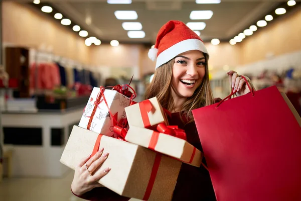 Happy girl shopping gifts in mall on christmas sale. New year holidays shopping idea concept. Smiling woman with colorful paper presents bags and gift boxes wearing christmas hat in store or shop. — Stock Photo, Image