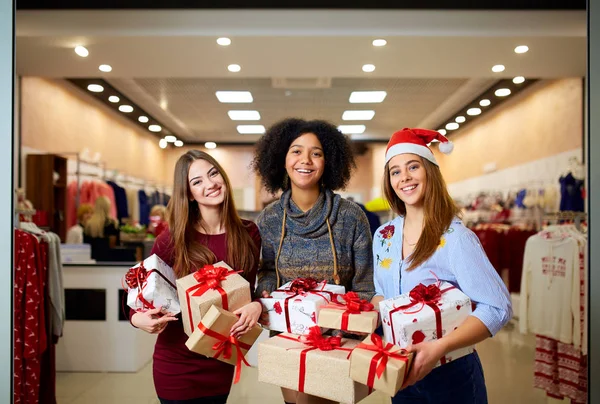 Three mixed race women with gift boxes in hands at store. Multi ethnic girls smiling with presents on christmas new year sale. Caucasian and african american females shopping presents for holidays. — Stock Photo, Image