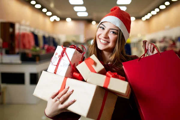 Happy girl shopping gifts in mall on christmas sale. New year holidays shopping idea concept. Smiling woman with colorful paper presents bags and gift boxes wearing christmas hat in store or shop. — Stock Photo, Image