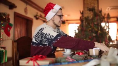 Oturma ve köknar ağacı evde yakınındaki bir hediye-yeni yıl için üzerinde bir yay bağlama adam sakallı. Dekorasyon çam dalları, koniler, baston şekerler ile kağıt, Noel şapka kaydırma mevcut giyen adam kutuları.