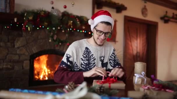 El hombre barbudo que se sienta y que ata el lazo a los regalos para Nuevo Año cerca de la chimenea. Tipo con sombrero de Navidad envolviendo cajas de regalo en papel añadiendo ramas de abeto, conos, caramelos de caña. Concepto de no masculinidad . — Vídeo de stock