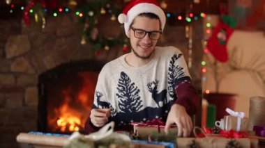 Oturma ve şömine yakınındaki bir hediye-yeni yıl için üzerinde bir yay bağlama adam sakallı. Noel şapka kaydırma mevcut giyen adam ekleme çam dalları, koniler, baston şekerler gazetede kutuları. Unmasculinity kavramı.