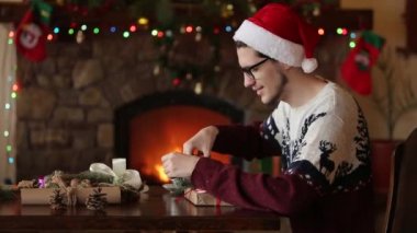 Oturan adam sakallı ve hediyeler Noel şapka kutuları kağıt, ambalaj şömine, yakınındaki yay bağlama dekorasyon çam dalları, koniler, baston şekerler, sunan gösterilen kameraya sunar.
