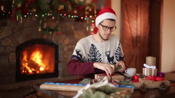 Bearded man sitting and tying a bow on a gifts for New Year near fireplace. Guy wearing Christmas hat wrapping present boxes in paper adding fir branches, cones, cane candies. Unmasculinity concept. — Stock Video