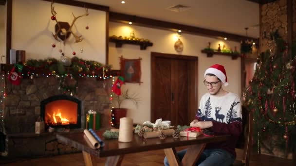 Skäggig man sitta och knyta en rosett på en gåvor för nyår nära eldstaden. Killen bär jul hatt inslagning nuvarande lådor i papper lägga till fir grenar, kottar, sockerrör godis. Unmasculinity koncept. — Stockvideo