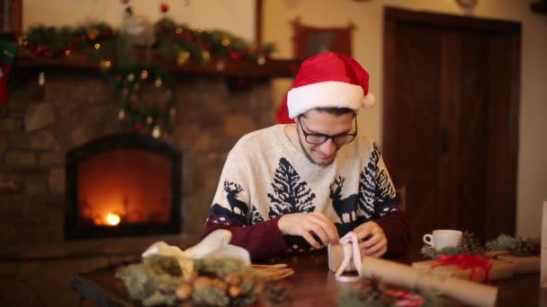 Heureux homme portant chapeaux santa ouverture boîte cadeau de Noël près de la cheminée avec guirlande clignotante. Un gars souriant a reçu un colis avec des cadeaux et profitant du déballage. Concept vacances d'hiver . — Video