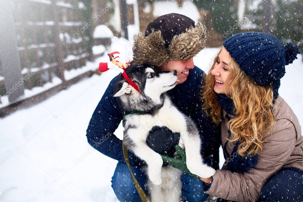Family portrait of cute happy couple hugging with their alaskan malamute dog licking mans face. Funny puppy wearing santa christmas deer antlers and licking mans face. Freedom lifestyle pet lovers.