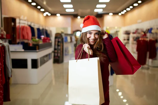 Happy girl shopping gifts in mall on christmas sale. New year holidays shopping idea concept. Smiling pretty caucasian woman with colorful paper presents bags wearing christmas hat in store or shop. — Stock Photo, Image