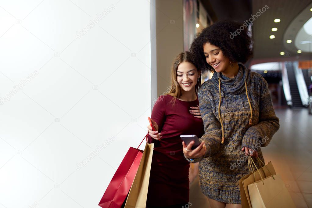 Two happy multiethnic young mixed race woman shopping for lingerie near white shop window copyspace, looking at smartphone screen. African american girl considers underwear with friend at shop front