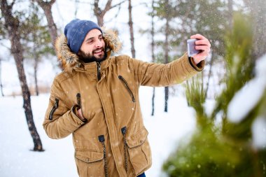 Akıllı telefon kamera ile fotoğraf gülümseyen sakallı adam giyer sıcak kış giysileri ve alma selfie. Yakışıklı adam mesaj ile cep telefonu ve apps içinde karlı ormanı kullanarak. Ormanda kar yağışı.