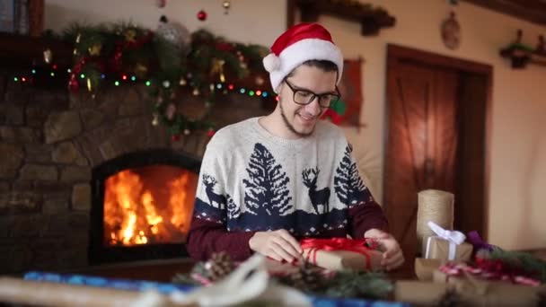 El hombre barbudo que se sienta y que ata el lazo a los regalos para Nuevo Año cerca de la chimenea. Tipo con sombrero de Navidad envolviendo cajas de regalo en papel añadiendo ramas de abeto, conos, caramelos de caña. Concepto de no masculinidad . — Vídeos de Stock