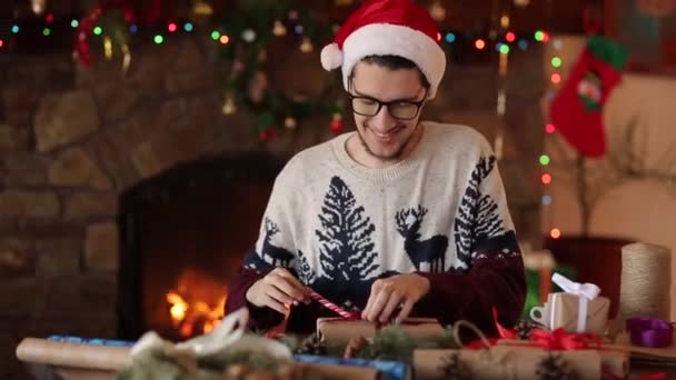 Бородатый мужчина сидит и вяжет бант на подарки на Новый год возле камина. Парень в рождественской шляпе, упаковывает подарочные коробки в бумагу, добавляет еловые ветки, шишки, тростниковые конфеты. Концепция немужественности . — стоковое видео