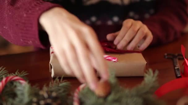 Vista cercana de las manos del hombre atando un lazo en un regalo para Navidad. Envoltura masculina cajas de regalo en papel añadiendo ramas de abeto, conos, caramelos de caña en la mesa de madera en casa. Vacaciones preparaciones . — Vídeo de stock