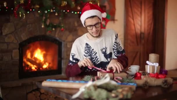 Bearded man sitting and tying a bow on a gifts near fireplace. Guy wearing Christmas hat wrapping boxes in paper, decorating with fir branches, cones, cane candies and then showing presents to camera. — Stock Video