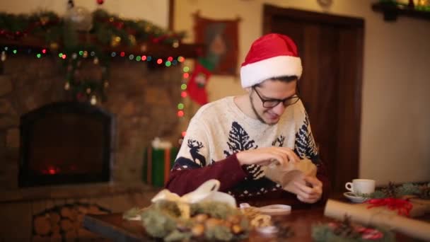 Счастливчик в шляпах Санты открывает рождественскую подарочную коробку возле камина с сверкающей гирляндой. Улыбающийся парень получил посылку с подарками и наслаждался распаковкой. Концепция зимнего отдыха . — стоковое видео