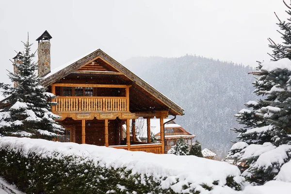 Christmas wooden mansion in mountains on snowfall winter day. Cozy chalet on ski resort near pine forest. Cottage of round timber with wooden balcony. Fir-trees covered with snow. Chimneys of stone. — Stock Photo, Image