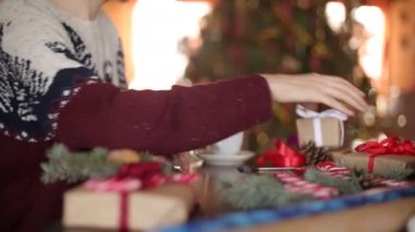 Yakın görünümünü bir hediyeler Noel için bir yay bağlama eller mans. Erkek mevcut kutuları ekleme çam dalları, koniler, cane kağıt kaydırma masada ahşap evde yılbaşı ağacı yakınındaki şekerler.