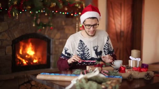 Bearded man sitting and tying a bow on a gifts for New Year near fireplace and looks to camera. Guy wearing Christmas hat wrapping present boxes in paper adding fir branches, cones, cane candies. — Stock Video