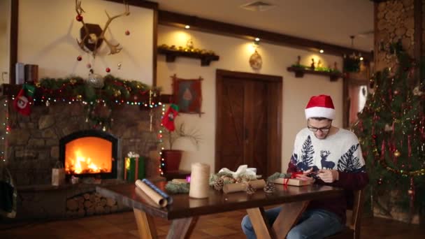 Bearded man sitting and tying bow on a gifts near fireplace, wearing Christmas hat wrapping boxes in paper, decorating with fir branches, cones, cane candies, then showing offering presents to camera. — Stock Video