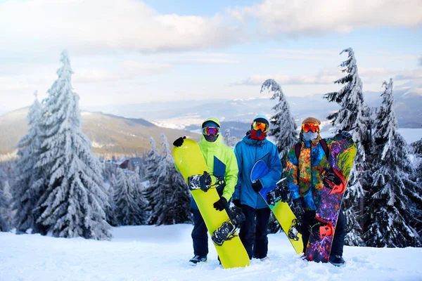Tres snowboarders posando en la estación de esquí. Amigos subiendo a la cima de la montaña llevando sus tablas de snowboard a través del bosque para freeride backcountry y usando gafas reflectantes, ropa de moda colorida . —  Fotos de Stock