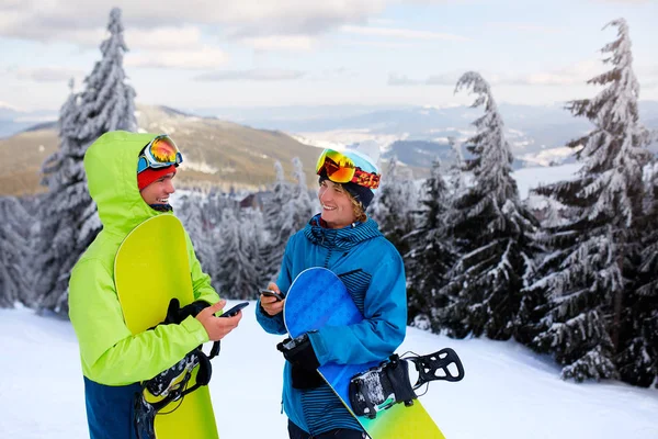 To snøbrettkjørere som snakker og holder smarttelefoner. Venner som ser på mobiler og kommuniserer på sosiale nettverk som deler sine bilder i app med internettilknytning iført fargerike motedrakter . – stockfoto