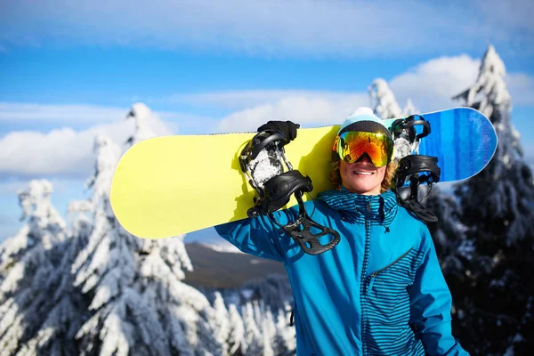Sonriente snowboarder posando llevando snowboard al hombro en la estación de esquí cerca del bosque antes de freeride backcountry y usando gafas reflectantes, colorido atuendo de moda. Equipo de snowboard moderno . —  Fotos de Stock