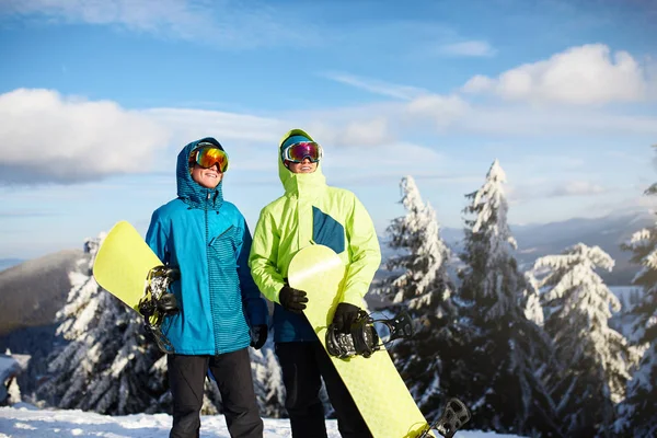Dos snowboarders posando en la estación de esquí. Jinetes amigos llevando sus tablas de snowboard a través del bosque para freeride backcountry y con gafas reflectantes, traje de moda de colores. Área del espacio de copia . —  Fotos de Stock
