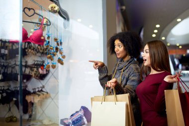 İki mutlu çok ırklı genç karışık ırk kadın iç çamaşırı giyim butik vitrin, yakın için alışveriş karar olup satın almak. Afro-Amerikan kız iç çamaşırı dükkanı açık adlı arkadaşıyla dikkate alır.