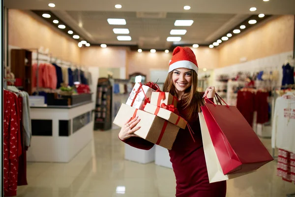 Happy girl shopping gifts in mall on christmas sale. New year holidays shopping idea concept. Smiling woman with colorful paper presents bags and gift boxes wearing christmas hat in store or shop. — Stock Photo, Image