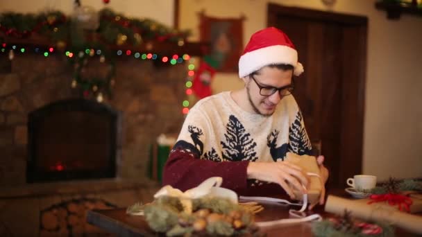 Hombre feliz con sombreros de santa apertura de la caja de regalo de Navidad cerca de la chimenea con guirnalda intermitente. El chico sonriente recibió un paquete con regalos y disfrutando desempacando el paquete. Concepto vacaciones de invierno . — Vídeos de Stock