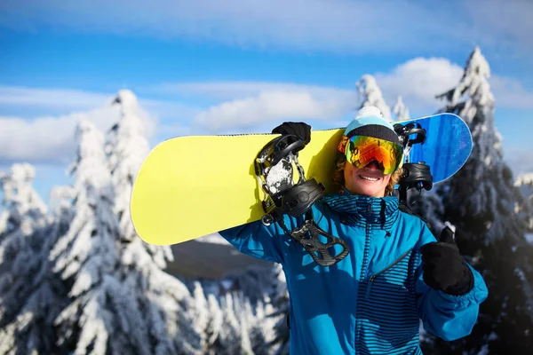 Smiling snowboarder posing carrying snowboard on shoulders at ski resort near forest before freeride session. Rider showing thumb up sign wearing polarized goggles. Modern snowboarding equipment. — Stock Photo, Image
