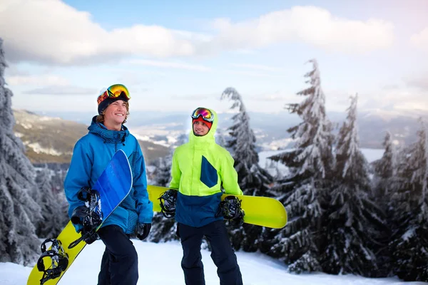 Dos snowboarders caminando en la estación de esquí. Amigos subiendo a la cima de la montaña llevando sus tablas de snowboard a través del bosque para freeride backcountry y usando gafas reflectantes, ropa de moda colorida . —  Fotos de Stock